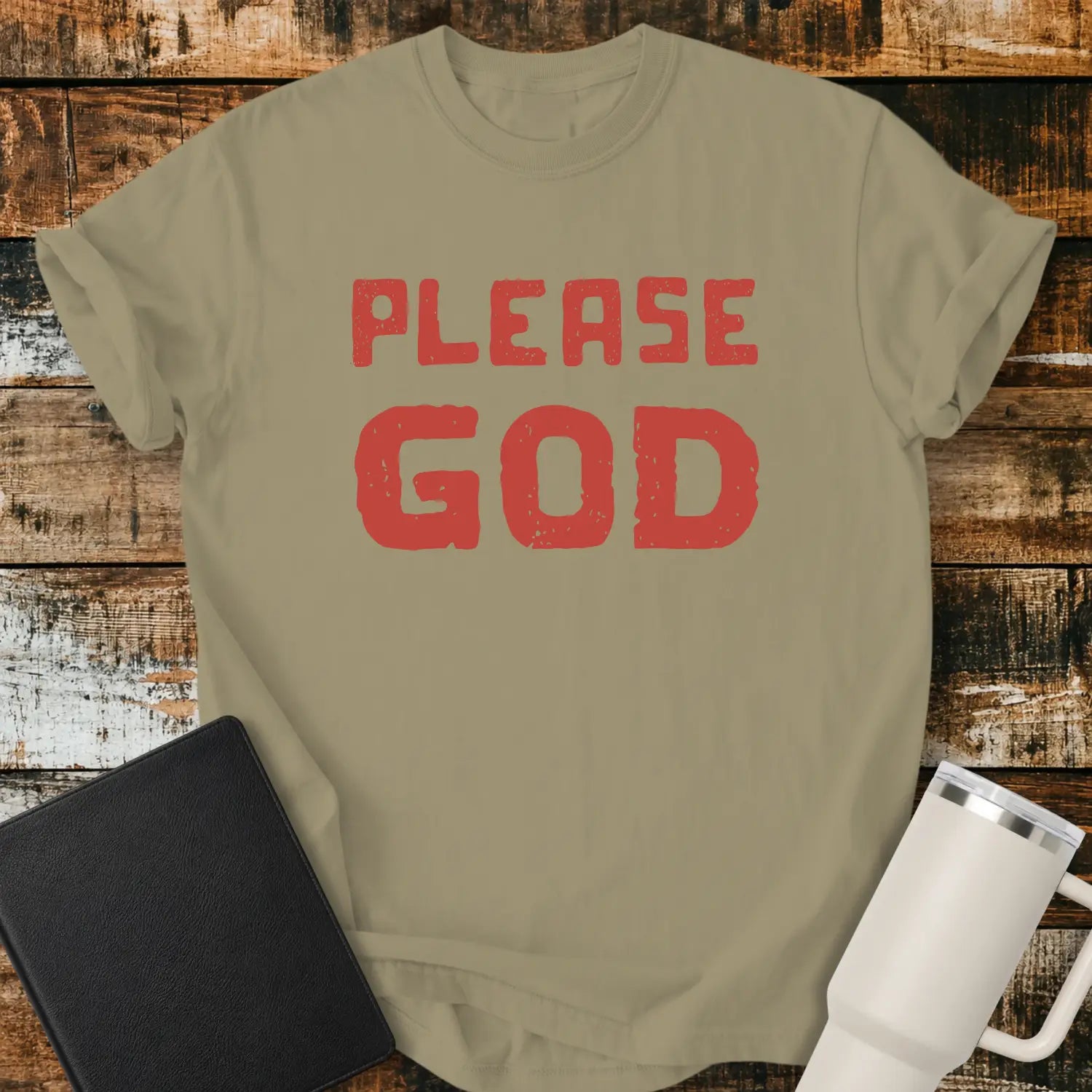 khaki T-shirt mockup featuring the simple phrase “PLEASE GOD” in large distressed red block lettering. The minimalist design is centered on the shirt, creating a bold faith-inspired statement. The shirt is displayed on a rustic wood plank background beside a black Bible and a white insulated tumbler.