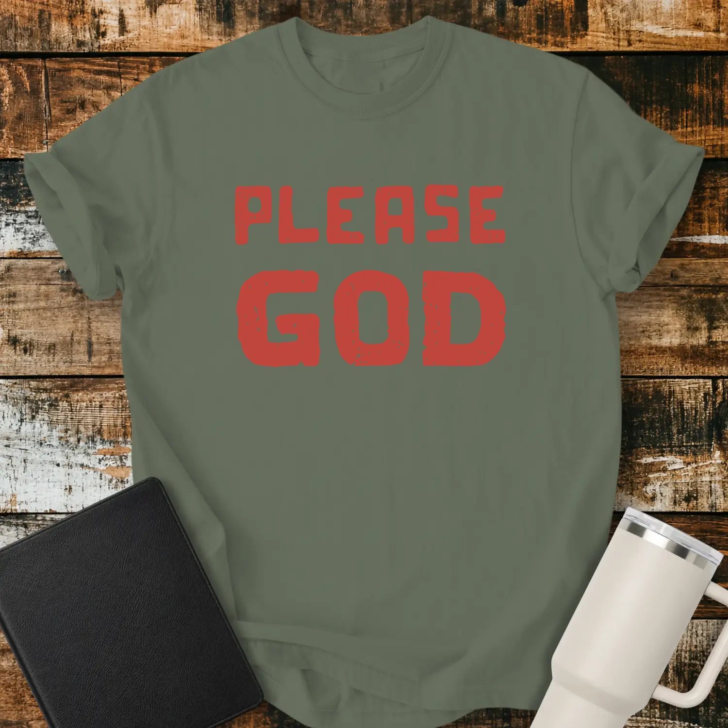 moss green T-shirt mockup featuring the simple phrase “PLEASE GOD” in large distressed red block lettering. The minimalist design is centered on the shirt, creating a bold faith-inspired statement. The shirt is displayed on a rustic wood plank background beside a black Bible and a white insulated tumbler.