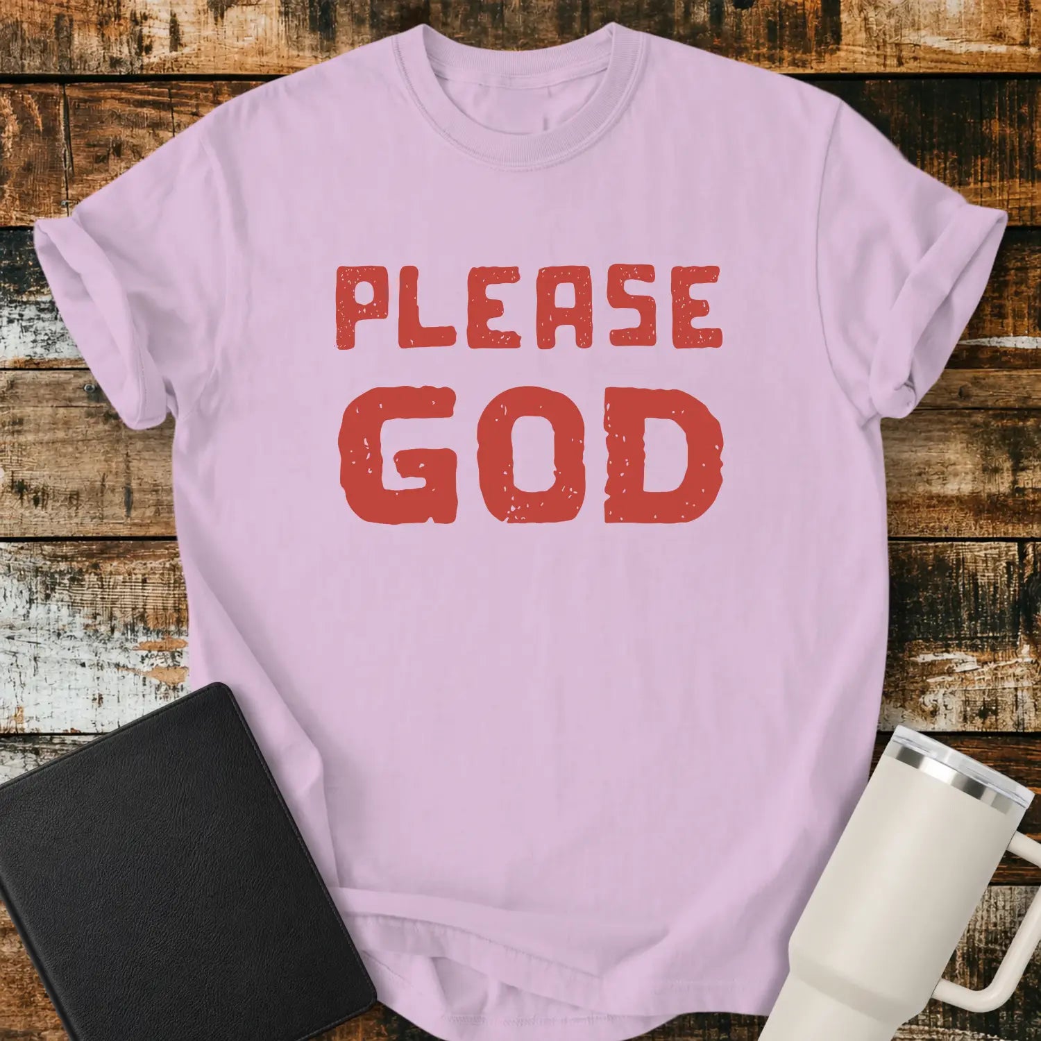 orchid purple T-shirt mockup featuring the simple phrase “PLEASE GOD” in large distressed red block lettering. The minimalist design is centered on the shirt, creating a bold faith-inspired statement. The shirt is displayed on a rustic wood plank background beside a black Bible and a white insulated tumbler.