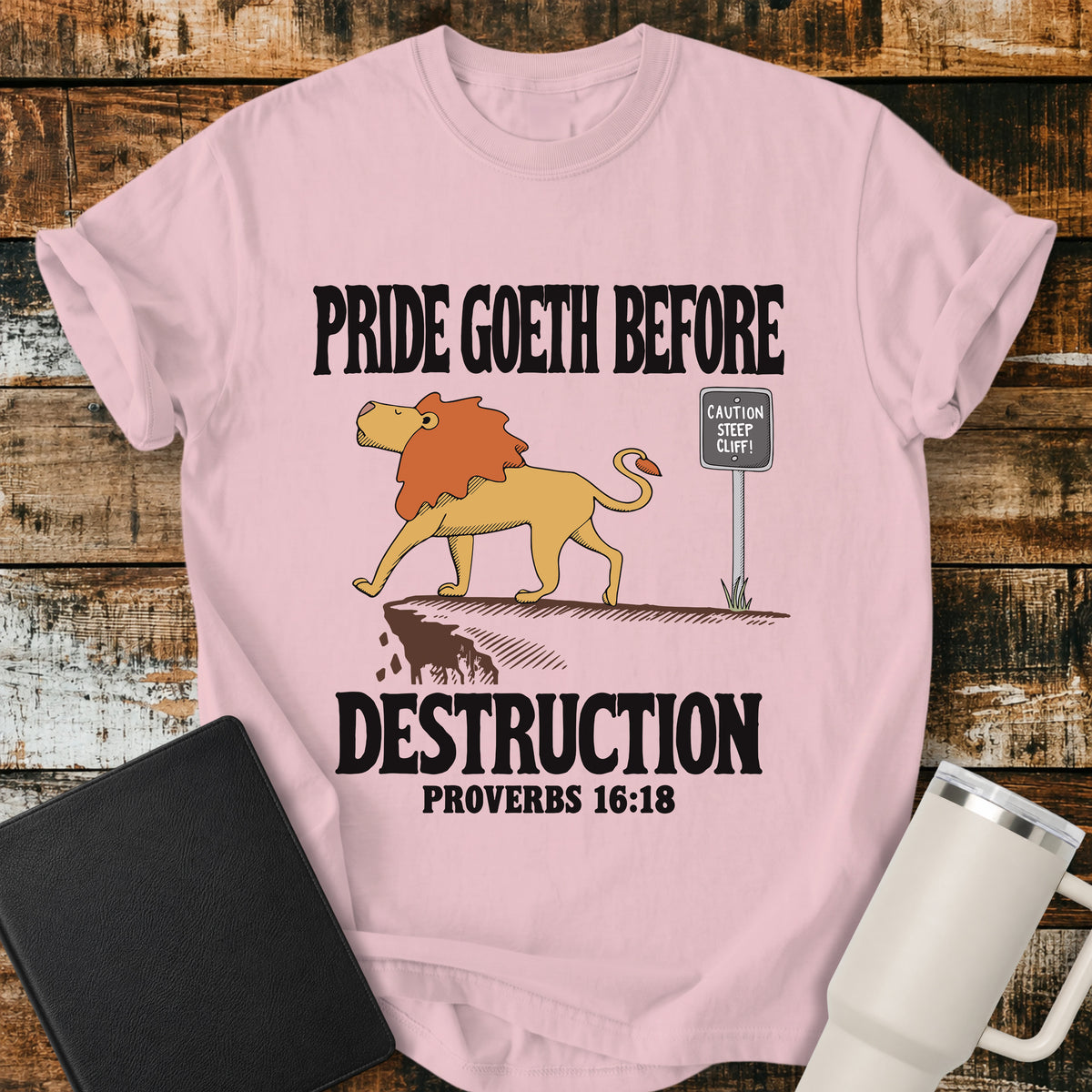blossom pink Christian t-shirt featuring a lion confidently approaching a steep cliff, with bold vintage lettering reading “Pride Goeth Before Destruction” and Proverbs 16:18.