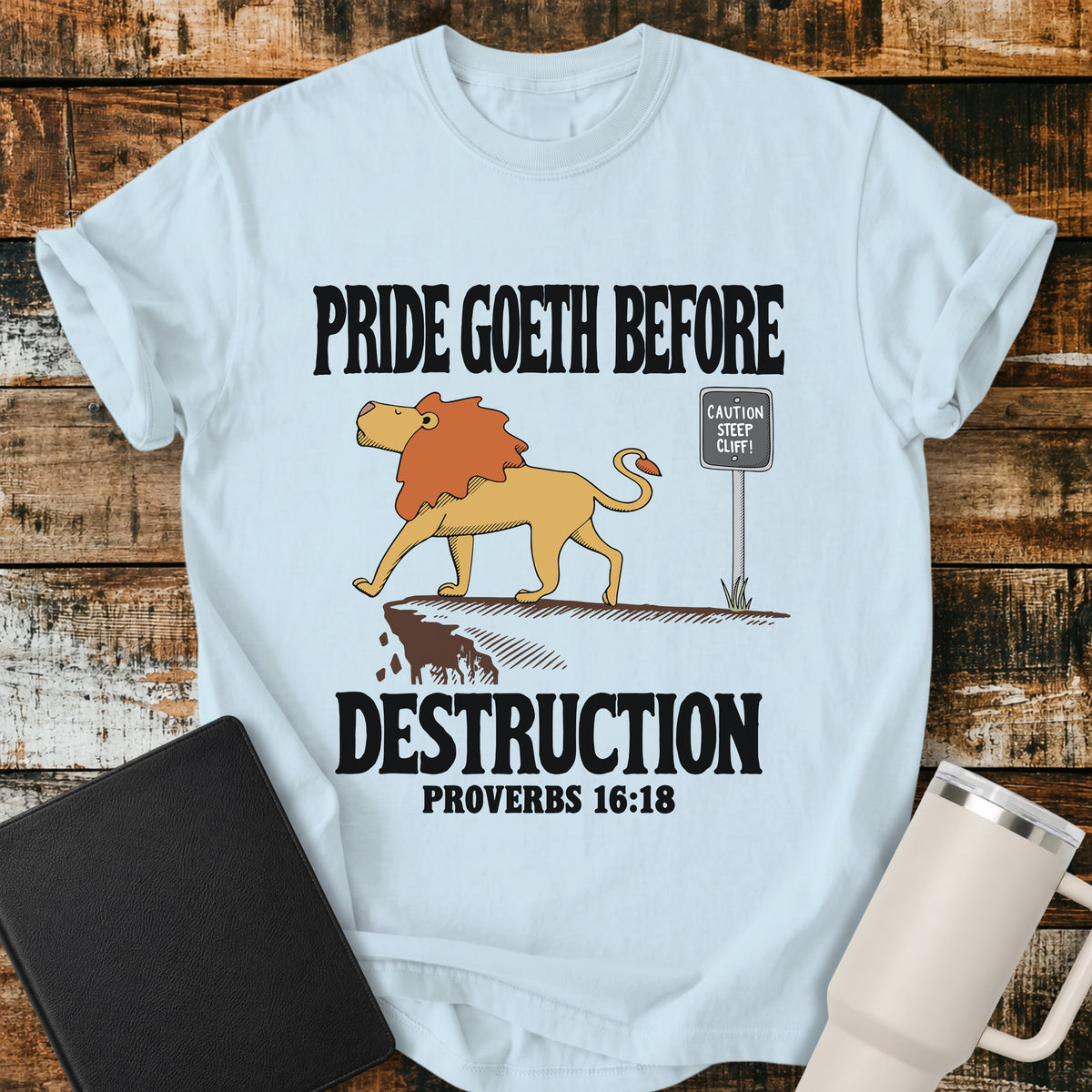 chambray blue Christian t-shirt featuring a lion confidently approaching a steep cliff, with bold vintage lettering reading “Pride Goeth Before Destruction” and Proverbs 16:18.