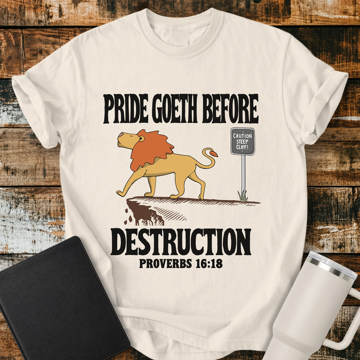 ivory Christian t-shirt featuring a lion confidently approaching a steep cliff, with bold vintage lettering reading “Pride Goeth Before Destruction” and Proverbs 16:18.