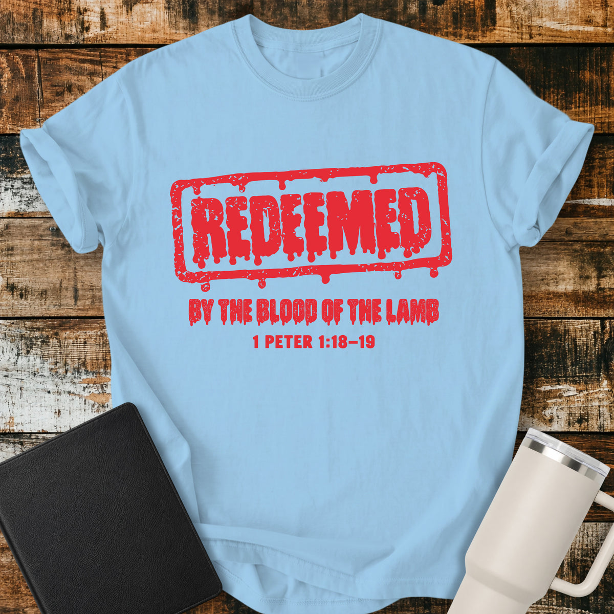 hydrangea blue Christian T-shirt featuring a bold red, distressed stamp-style “REDEEMED” graphic with dripping details, paired with “BY THE BLOOD OF THE LAMB” and the Bible reference “1 PETER 1:18–19.”