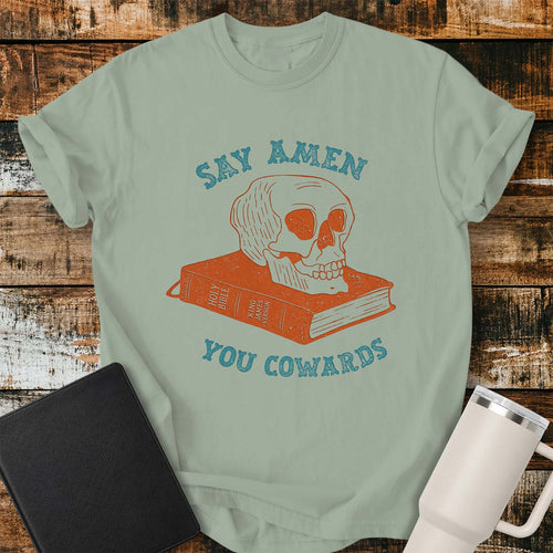 bay green shirt featuring a vintage-inspired illustration of a skull resting on a closed King James Bible. Distressed blue lettering above and below reads, “SAY AMEN” and “YOU COWARDS.” The artwork uses muted rust and blue tones for a retro look. The shirt is displayed on a rustic wood plank background with a black Bible on the left and a white insulated tumbler on the right.