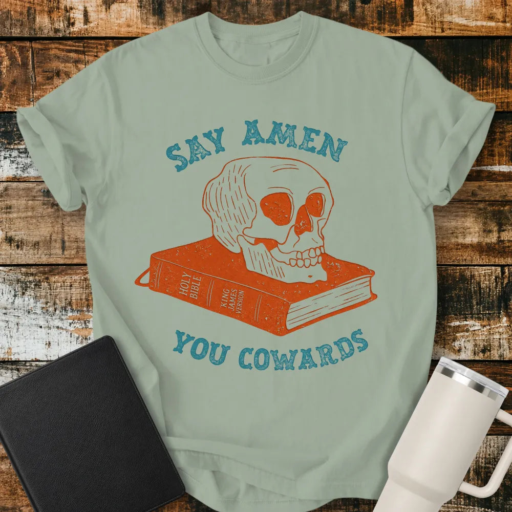 bay green graphic T-shirt mockup featuring a distressed skull resting on a King James Version Holy Bible with the phrase “Say Amen You Cowards” in vintage-style blue lettering, styled with a black Bible and white tumbler on a rustic wood background.