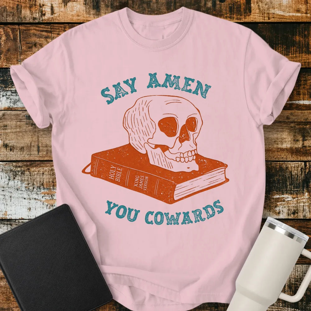 blossom pink graphic T-shirt mockup featuring a distressed skull resting on a King James Version Holy Bible with the phrase “Say Amen You Cowards” in vintage-style blue lettering, styled with a black Bible and white tumbler on a rustic wood background.