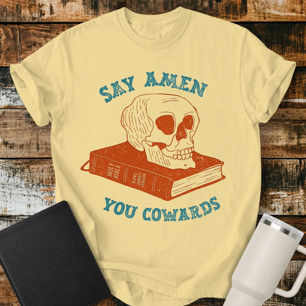 butter yellow graphic T-shirt mockup featuring a distressed skull resting on a King James Version Holy Bible with the phrase “Say Amen You Cowards” in vintage-style blue lettering, styled with a black Bible and white tumbler on a rustic wood background.