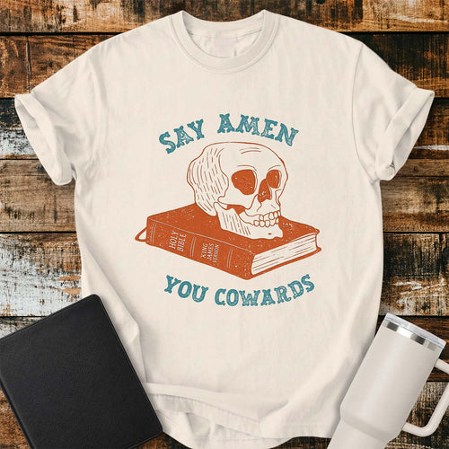 Ivory shirt featuring a vintage-inspired illustration of a skull resting on a closed King James Bible. Distressed blue lettering above and below reads, “SAY AMEN” and “YOU COWARDS.” The artwork uses muted rust and blue tones for a retro look. The shirt is displayed on a rustic wood plank background with a black Bible on the left and a white insulated tumbler on the right.