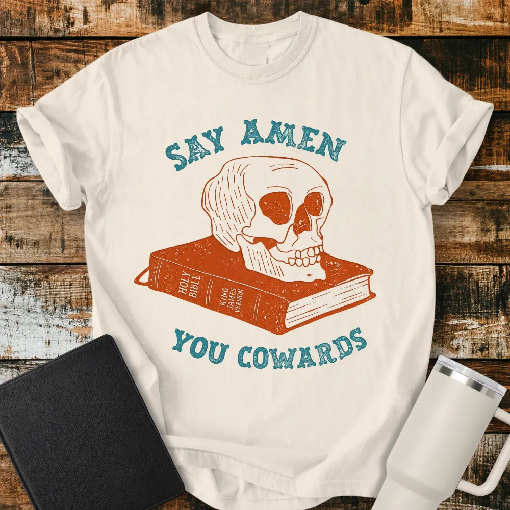Ivory graphic T-shirt mockup featuring a distressed skull resting on a King James Version Holy Bible with the phrase “Say Amen You Cowards” in vintage-style blue lettering, styled with a black Bible and white tumbler on a rustic wood background.