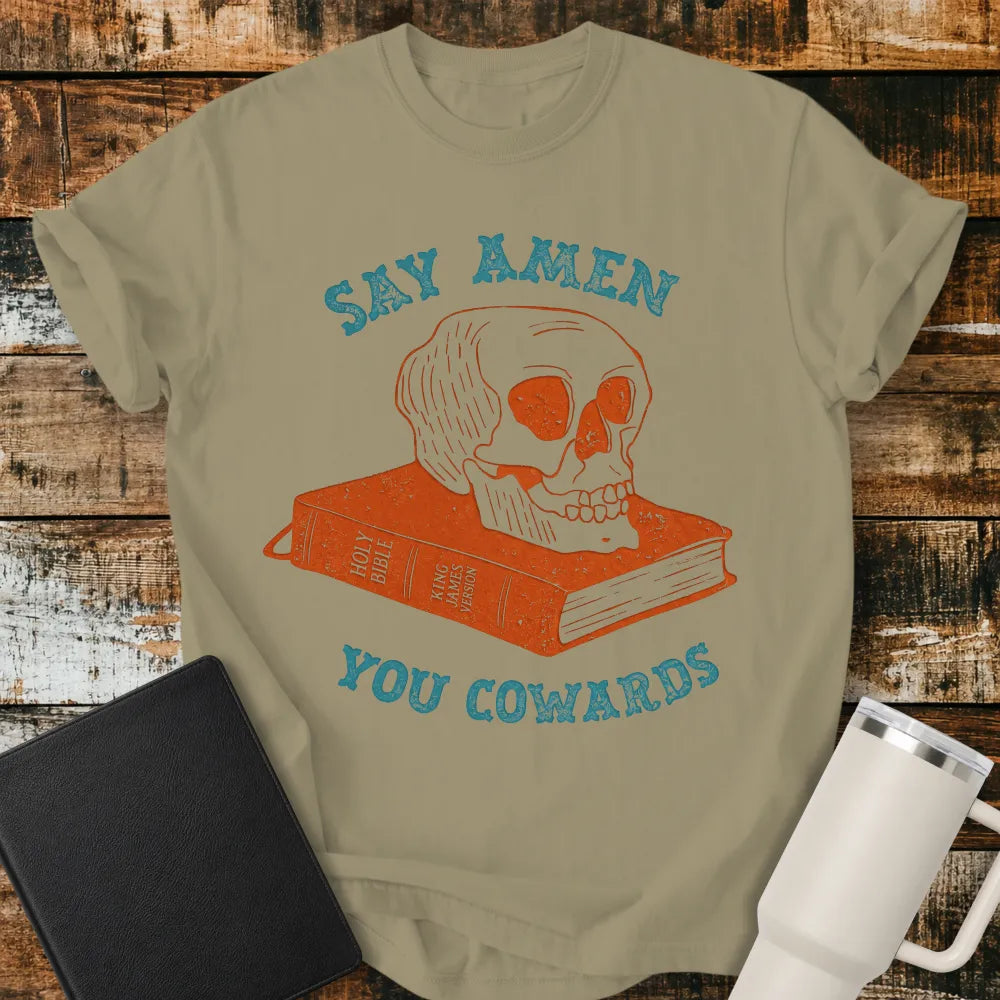 khaki brown graphic T-shirt mockup featuring a distressed skull resting on a King James Version Holy Bible with the phrase “Say Amen You Cowards” in vintage-style blue lettering, styled with a black Bible and white tumbler on a rustic wood background.