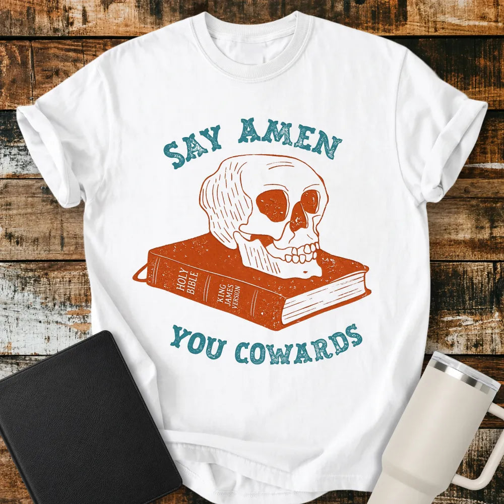 white graphic T-shirt mockup featuring a distressed skull resting on a King James Version Holy Bible with the phrase “Say Amen You Cowards” in vintage-style blue lettering, styled with a black Bible and white tumbler on a rustic wood background.