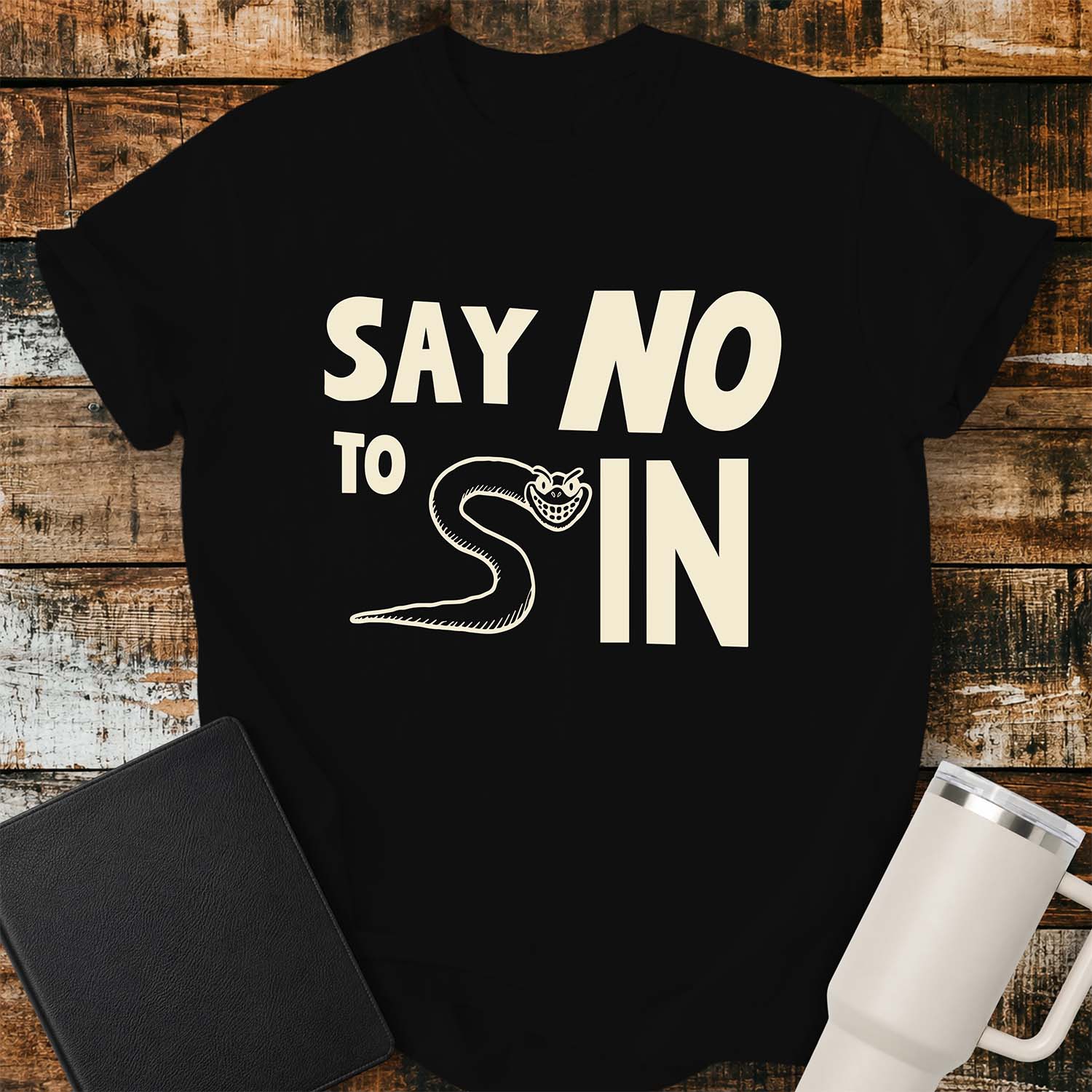 black T-shirt featuring bold black typography reading “SAY NO TO SIN,” with a smiling cartoon snake cleverly forming the letter “S” in “SIN.” The shirt is displayed on a rustic wood plank background alongside a black Bible and a white insulated tumbler.