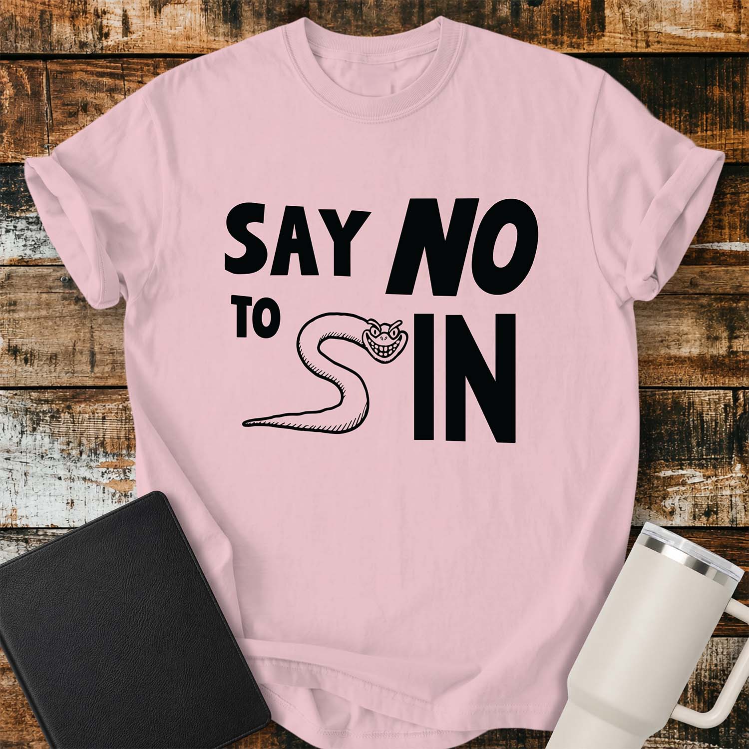 blossom pink T-shirt featuring bold black typography reading “SAY NO TO SIN,” with a smiling cartoon snake cleverly forming the letter “S” in “SIN.” The shirt is displayed on a rustic wood plank background alongside a black Bible and a white insulated tumbler.