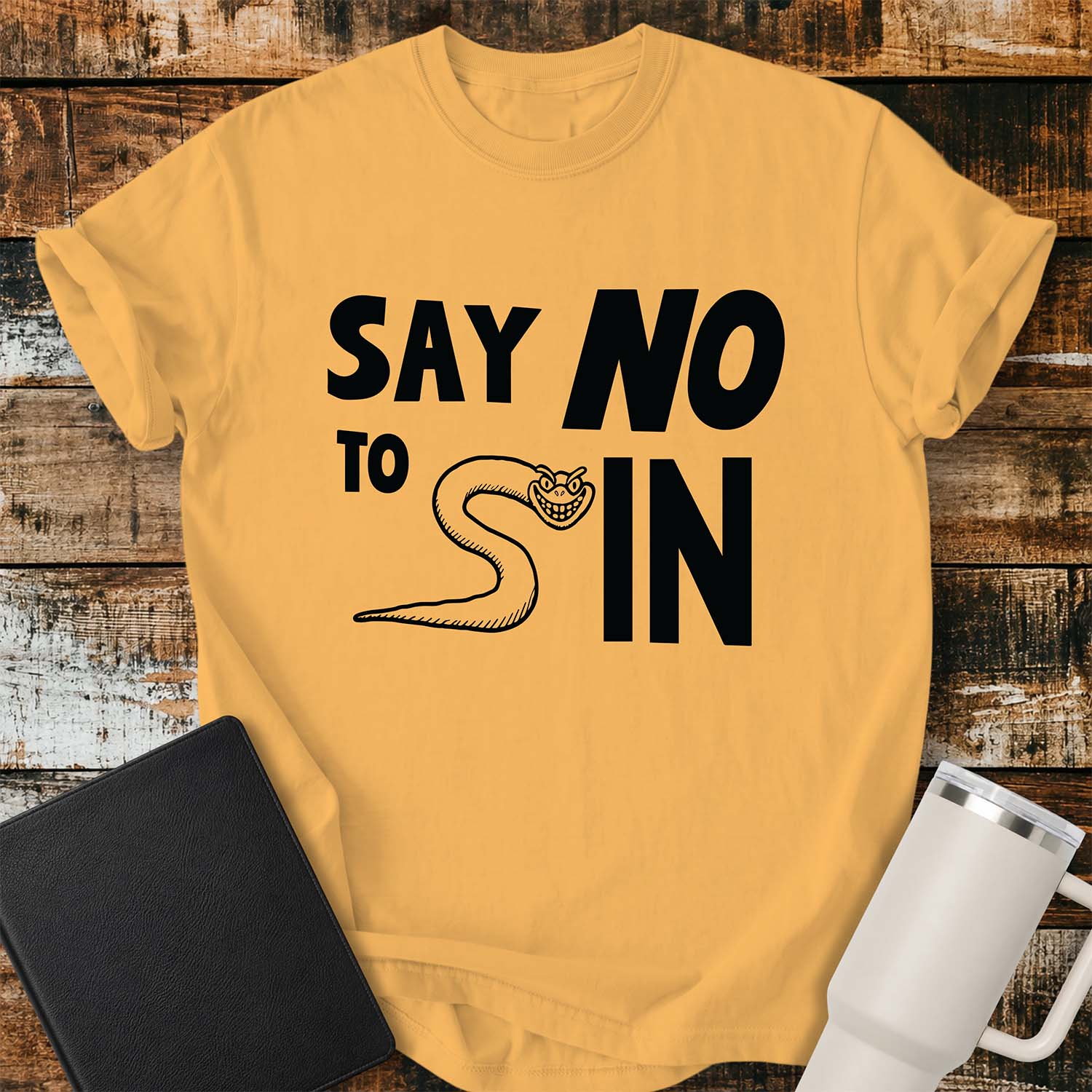 Citrus yellow T-shirt featuring bold black typography reading “SAY NO TO SIN,” with a smiling cartoon snake cleverly forming the letter “S” in “SIN.” The shirt is displayed on a rustic wood plank background alongside a black Bible and a white insulated tumbler.