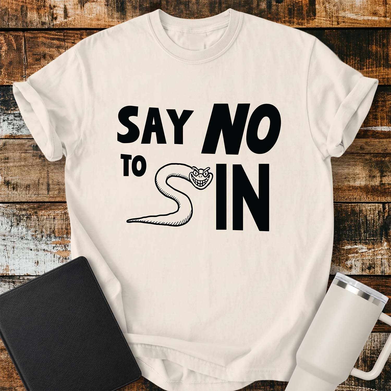 ivory T-shirt featuring bold black typography reading “SAY NO TO SIN,” with a smiling cartoon snake cleverly forming the letter “S” in “SIN.” The shirt is displayed on a rustic wood plank background alongside a black Bible and a white insulated tumbler.
