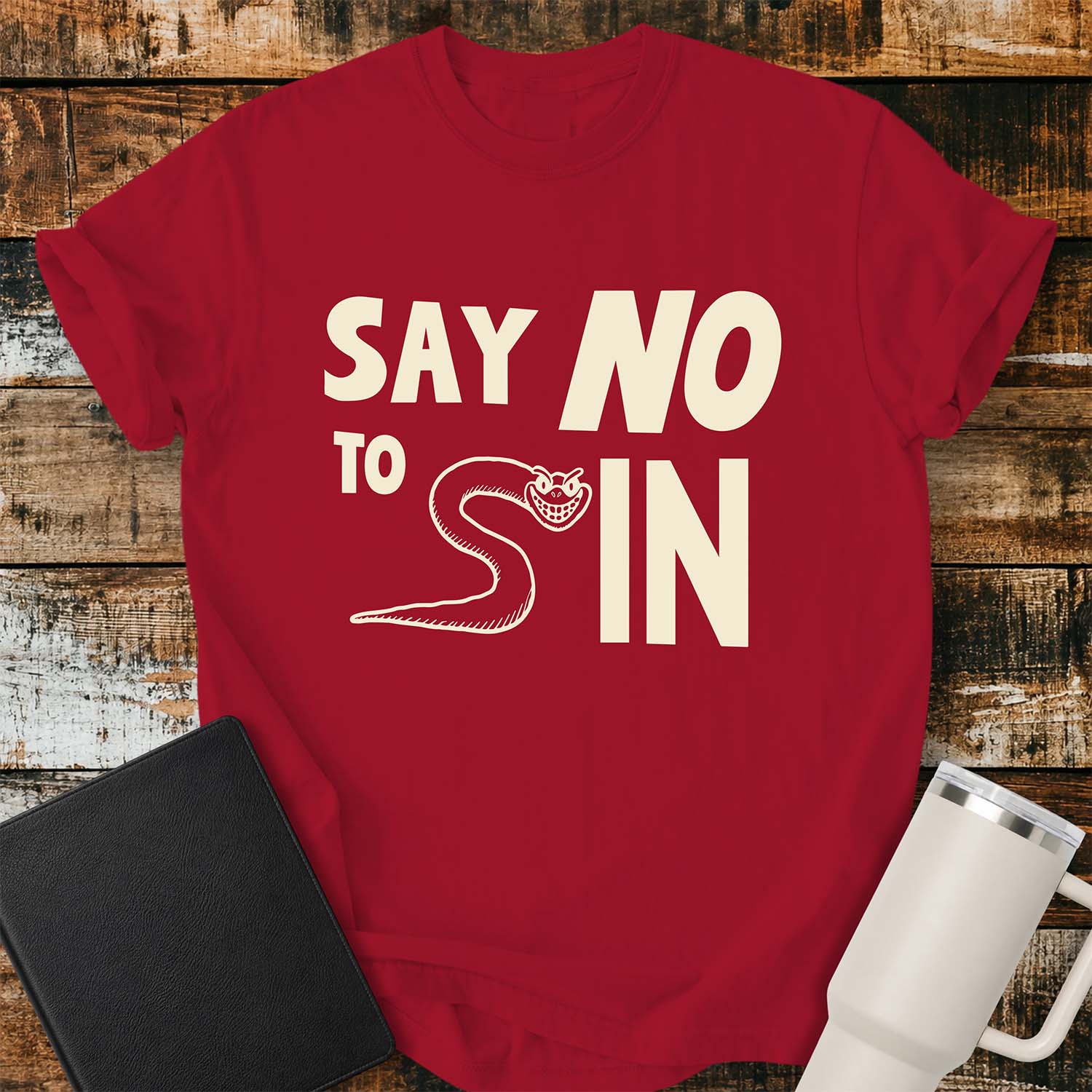 red T-shirt featuring bold black typography reading “SAY NO TO SIN,” with a smiling cartoon snake cleverly forming the letter “S” in “SIN.” The shirt is displayed on a rustic wood plank background alongside a black Bible and a white insulated tumbler.