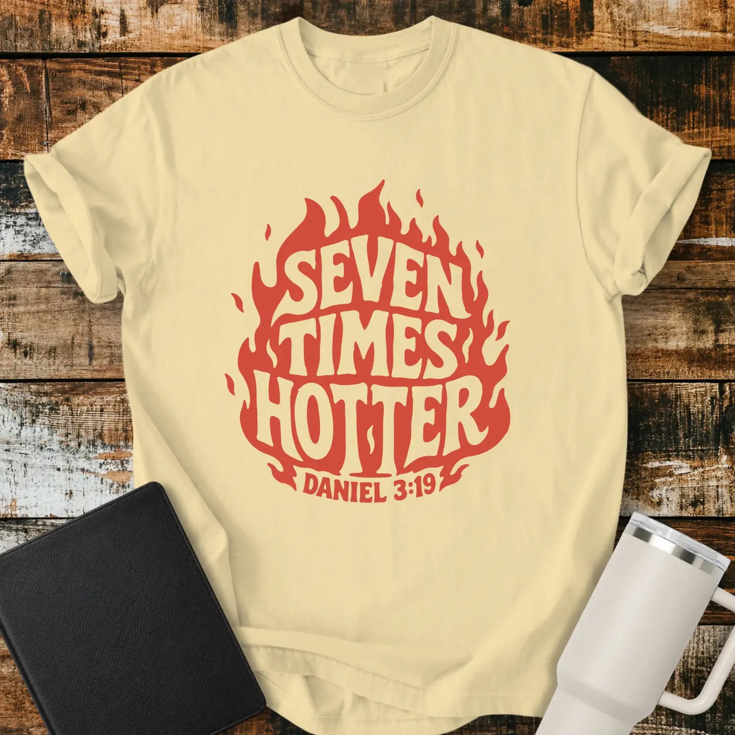 banana yellow T-shirt mockup featuring a retro red flame graphic with the words “SEVEN TIMES HOTTER” integrated into the flame design and “DANIEL 3:19” below. The shirt is displayed on a rustic wood background alongside a black Bible and a white insulated tumbler.