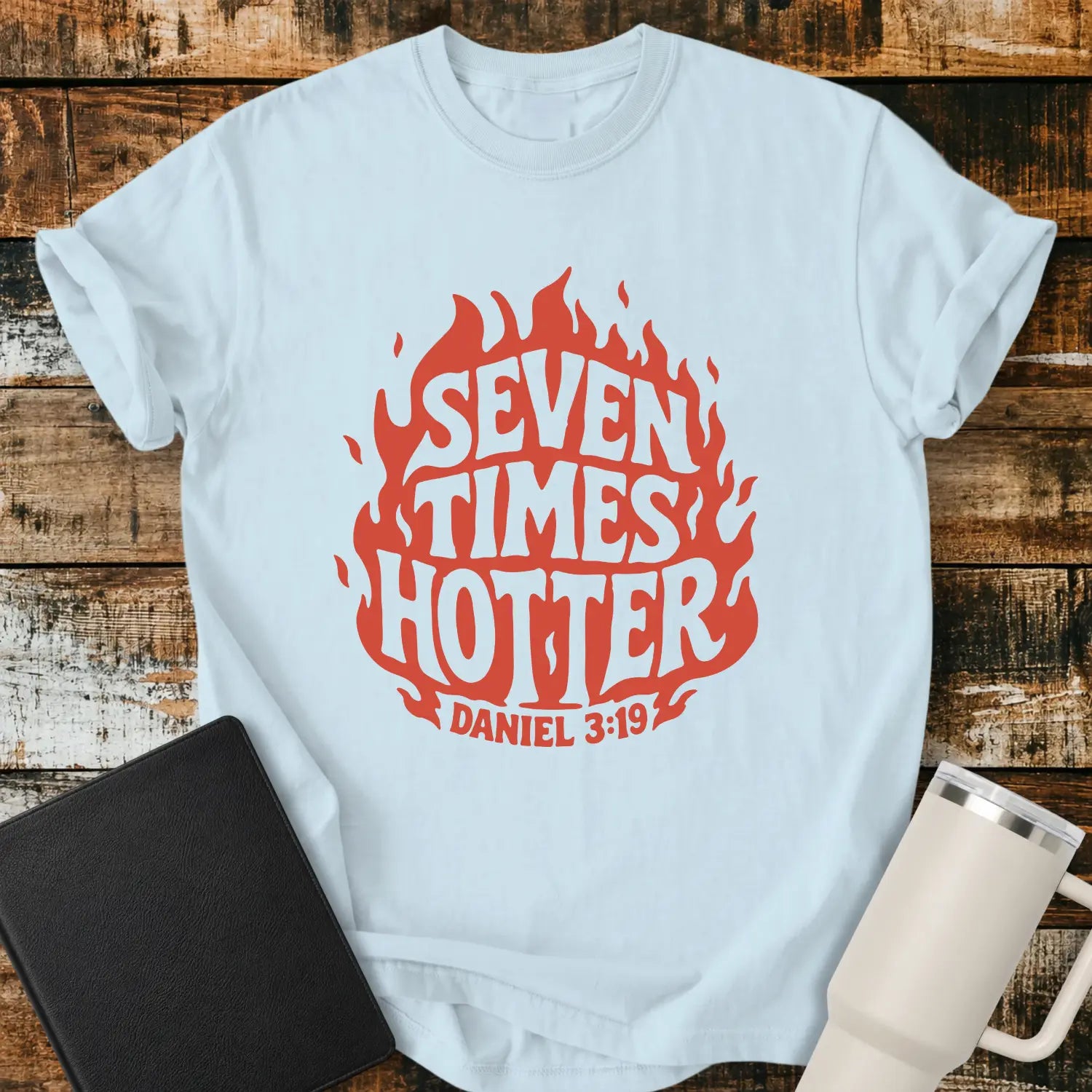 chambray blue T-shirt mockup featuring a retro red flame graphic with the words “SEVEN TIMES HOTTER” integrated into the flame design and “DANIEL 3:19” below. The shirt is displayed on a rustic wood background alongside a black Bible and a white insulated tumbler.