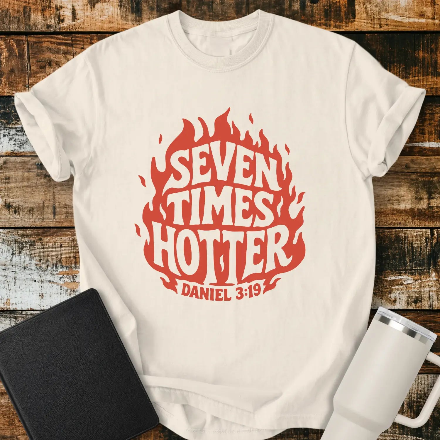 Ivory T-shirt mockup featuring a retro red flame graphic with the words “SEVEN TIMES HOTTER” integrated into the flame design and “DANIEL 3:19” below. The shirt is displayed on a rustic wood background alongside a black Bible and a white insulated tumbler.