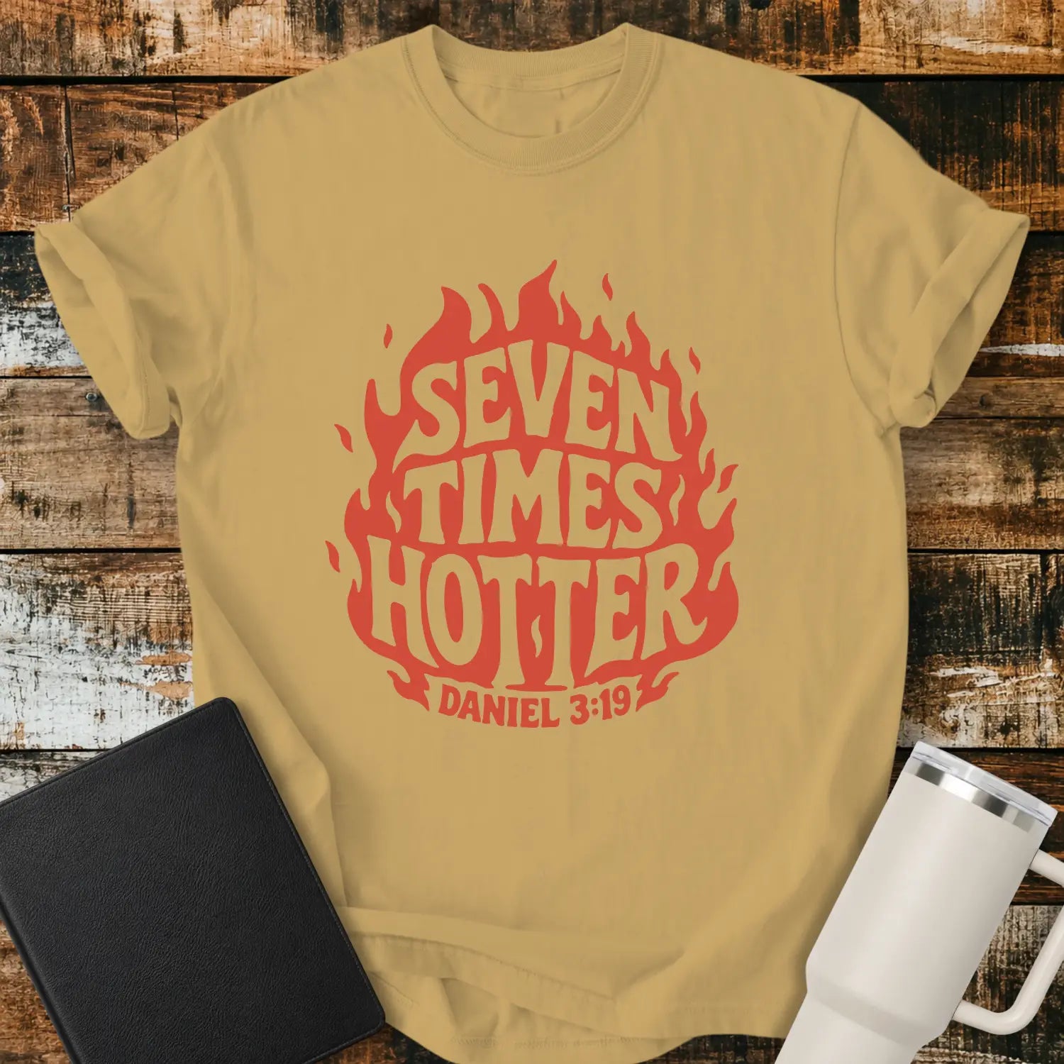 mustard yellow T-shirt mockup featuring a retro red flame graphic with the words “SEVEN TIMES HOTTER” integrated into the flame design and “DANIEL 3:19” below. The shirt is displayed on a rustic wood background alongside a black Bible and a white insulated tumbler.
