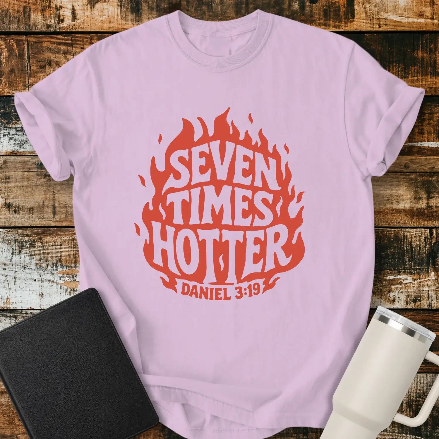 orchid purple T-shirt mockup featuring a retro red flame graphic with the words “SEVEN TIMES HOTTER” integrated into the flame design and “DANIEL 3:19” below. The shirt is displayed on a rustic wood background alongside a black Bible and a white insulated tumbler.
