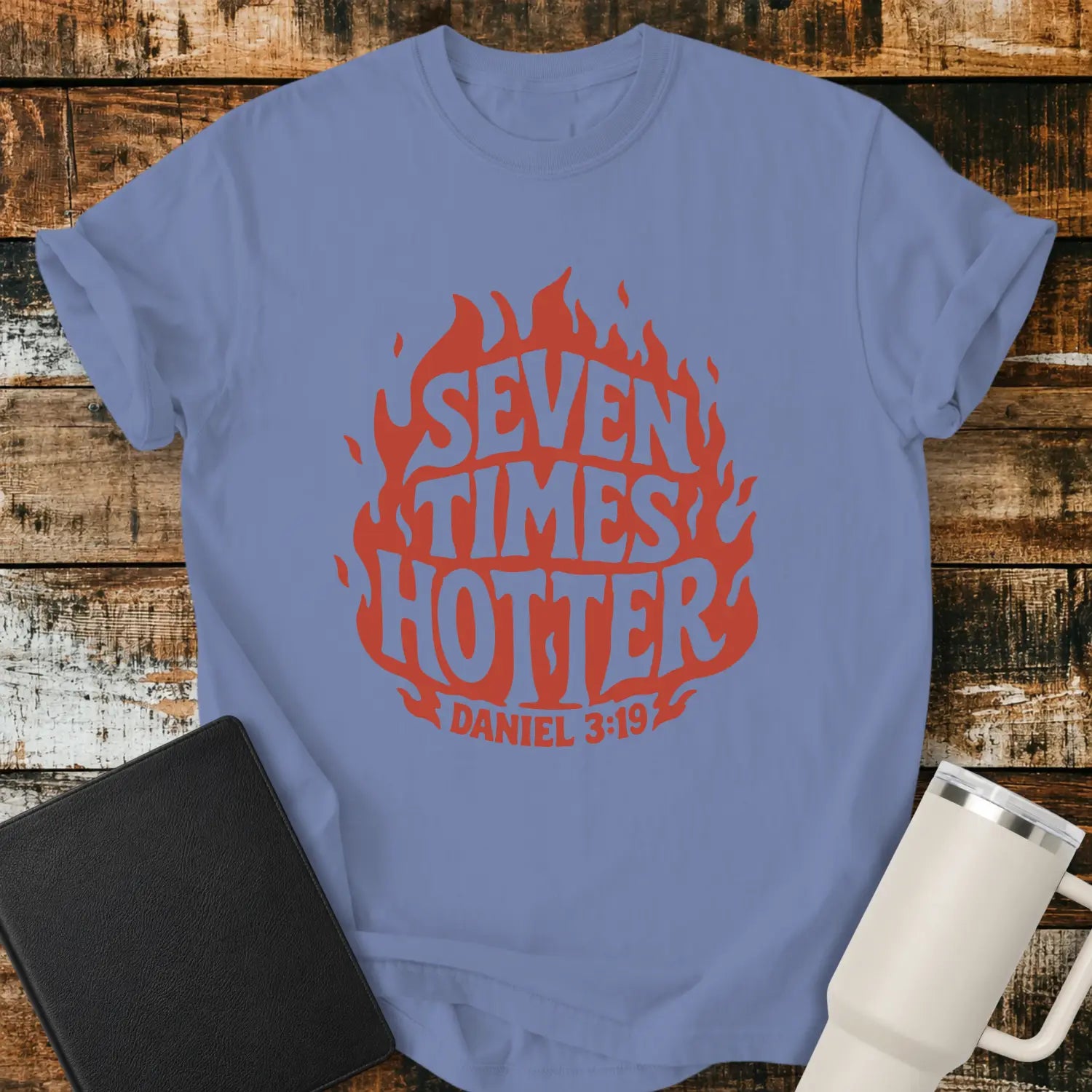 washed denim blue T-shirt mockup featuring a retro red flame graphic with the words “SEVEN TIMES HOTTER” integrated into the flame design and “DANIEL 3:19” below. The shirt is displayed on a rustic wood background alongside a black Bible and a white insulated tumbler.