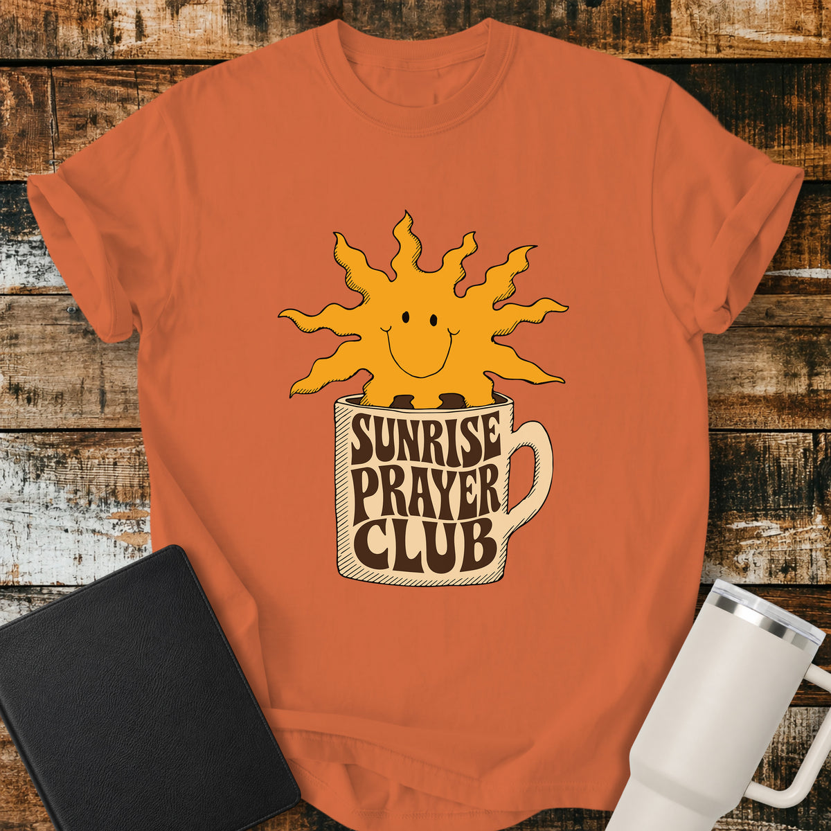 burnt orange flat-lay t-shirt mockup featuring a cheerful smiling sun rising from a coffee mug with the words "SUNRISE PRAYER CLUB" incorporated into the mug's retro lettering. The warm vintage-inspired design is centered on the shirt and displayed on a rustic wood plank background alongside a black leather Bible and a white insulated travel mug.