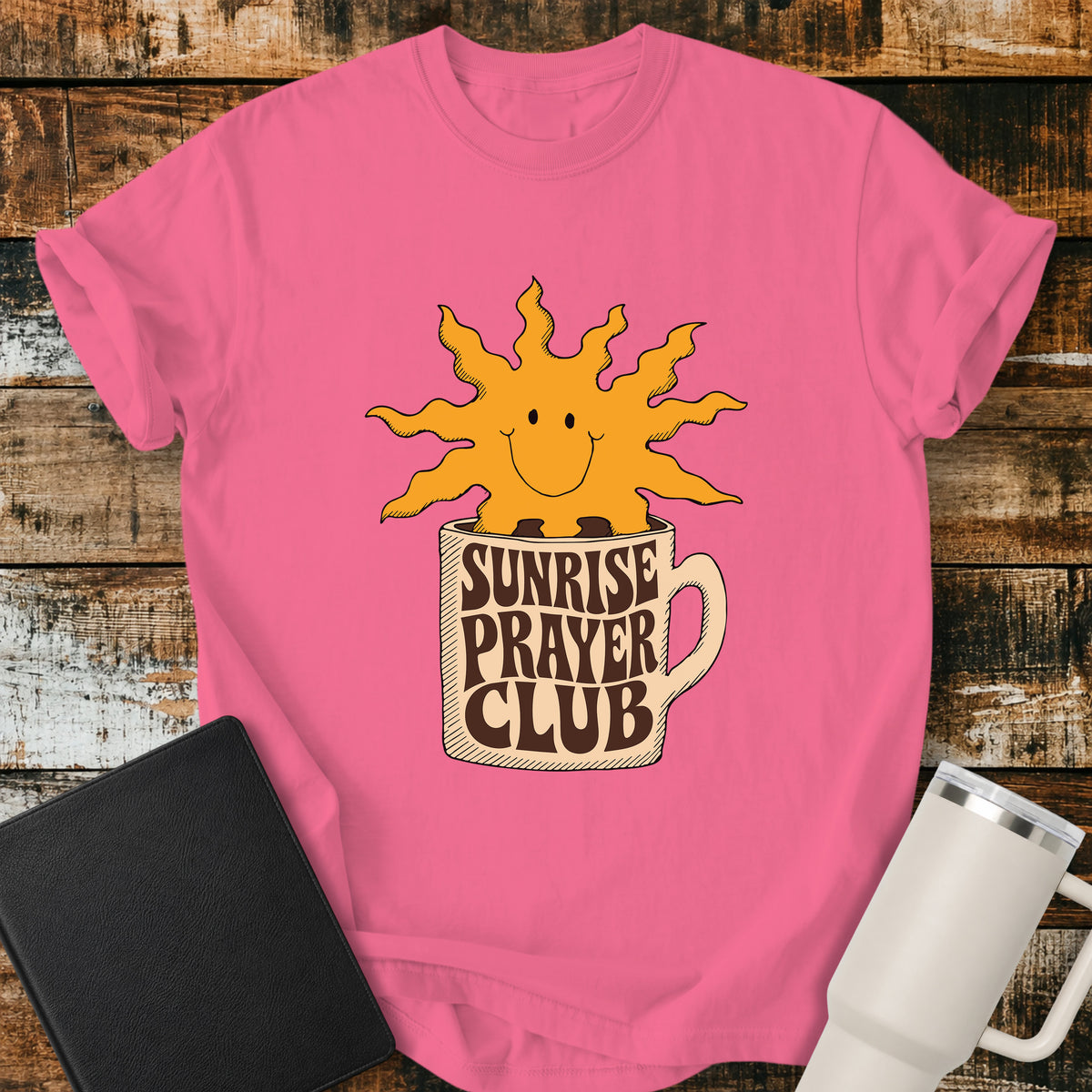 crunchberry pink flat-lay t-shirt mockup featuring a cheerful smiling sun rising from a coffee mug with the words "SUNRISE PRAYER CLUB" incorporated into the mug's retro lettering. The warm vintage-inspired design is centered on the shirt and displayed on a rustic wood plank background alongside a black leather Bible and a white insulated travel mug.