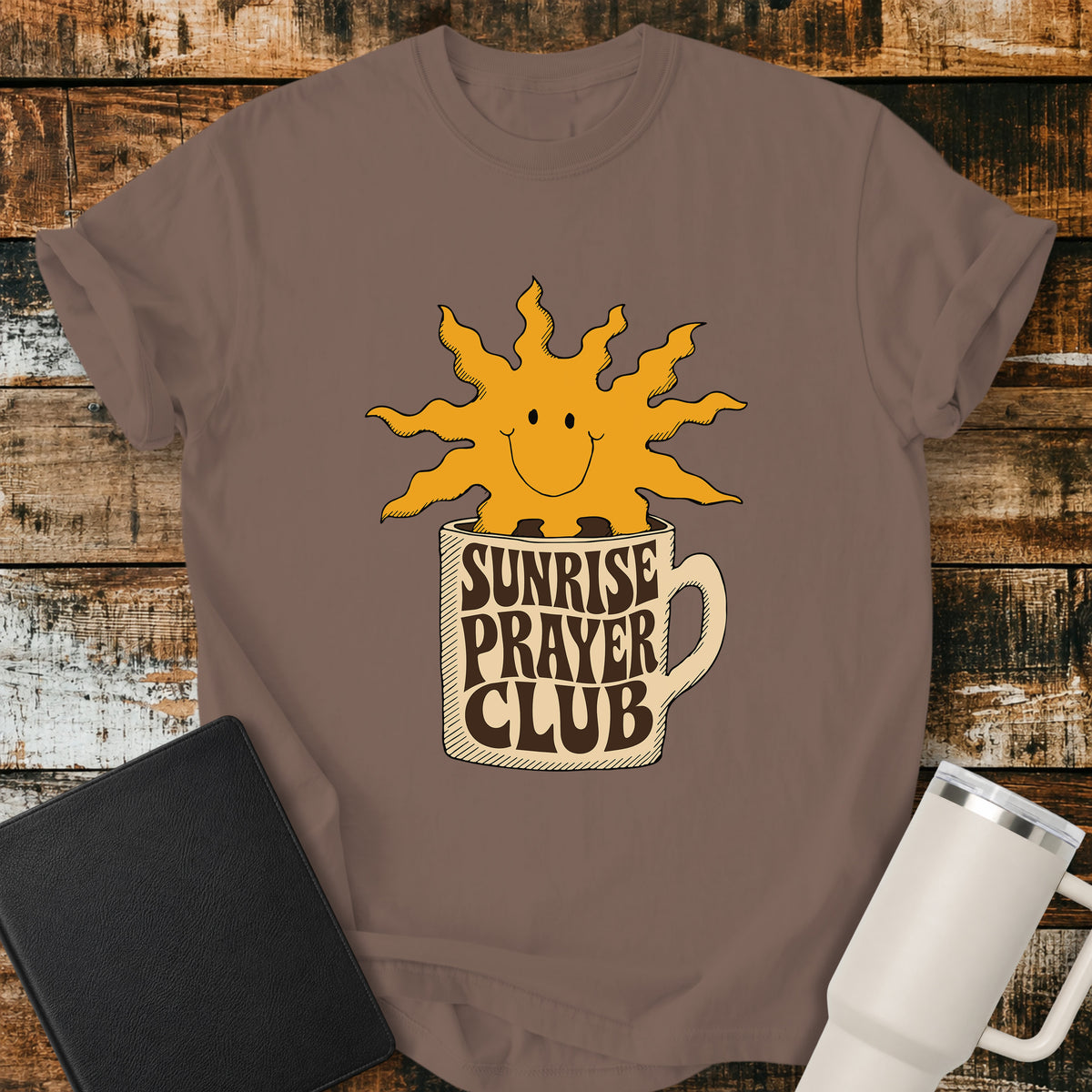 Espresso brown flat-lay t-shirt mockup featuring a cheerful smiling sun rising from a coffee mug with the words "SUNRISE PRAYER CLUB" incorporated into the mug's retro lettering. The warm vintage-inspired design is centered on the shirt and displayed on a rustic wood plank background alongside a black leather Bible and a white insulated travel mug.
