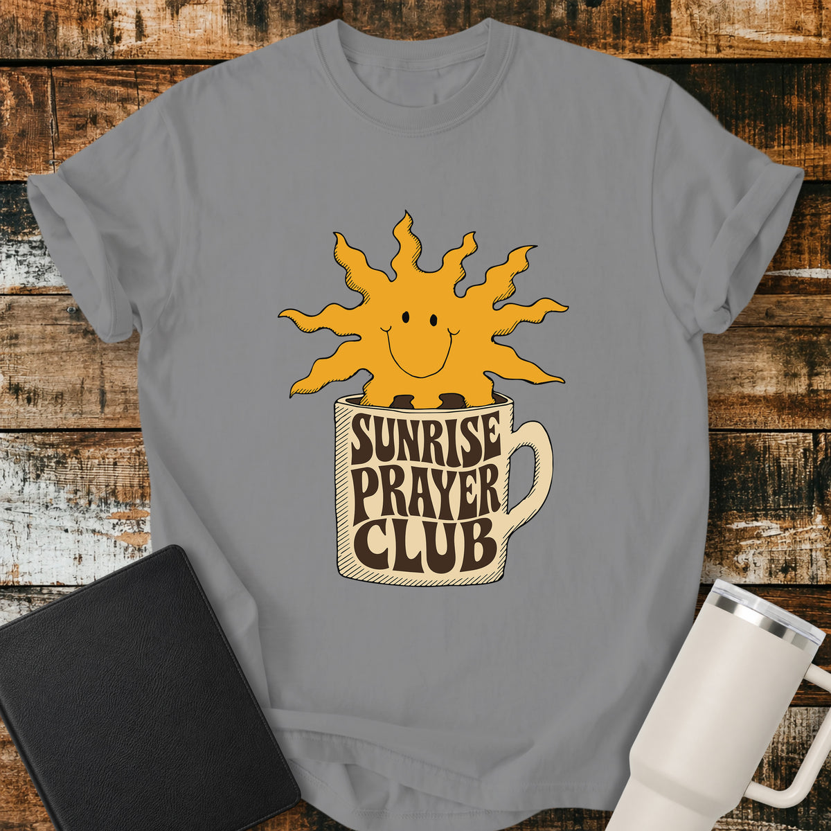 grey flat-lay t-shirt mockup featuring a cheerful smiling sun rising from a coffee mug with the words "SUNRISE PRAYER CLUB" incorporated into the mug's retro lettering. The warm vintage-inspired design is centered on the shirt and displayed on a rustic wood plank background alongside a black leather Bible and a white insulated travel mug.