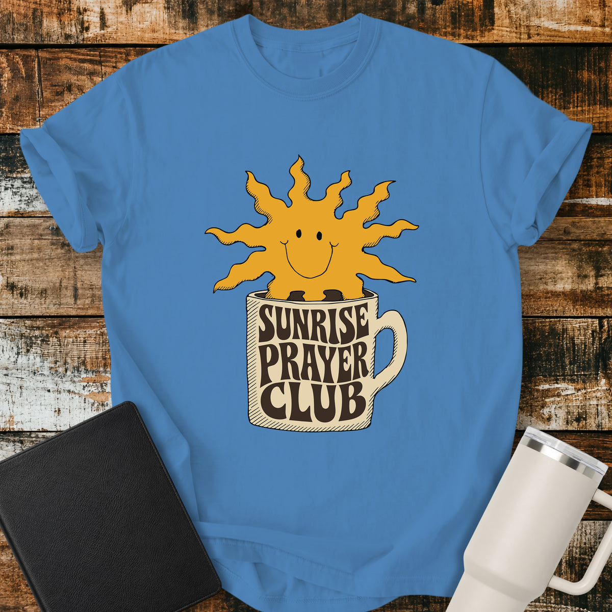 royal caribe blue flat-lay t-shirt mockup featuring a cheerful smiling sun rising from a coffee mug with the words "SUNRISE PRAYER CLUB" incorporated into the mug's retro lettering. The warm vintage-inspired design is centered on the shirt and displayed on a rustic wood plank background alongside a black leather Bible and a white insulated travel mug.