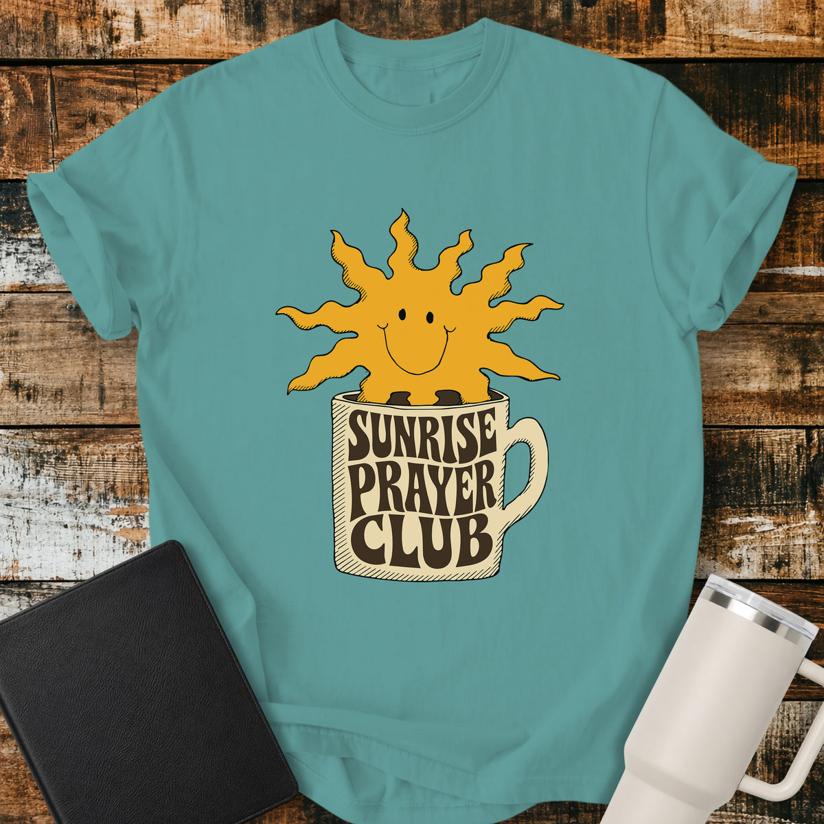 seafoam green flat-lay t-shirt mockup featuring a cheerful smiling sun rising from a coffee mug with the words "SUNRISE PRAYER CLUB" incorporated into the mug's retro lettering. The warm vintage-inspired design is centered on the shirt and displayed on a rustic wood plank background alongside a black leather Bible and a white insulated travel mug.