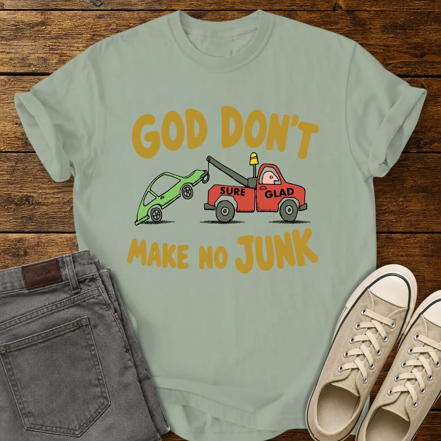 bay green T-shirt featuring a humorous cartoon of a red tow truck labeled “SURE GLAD” towing a broken green car beneath the phrase “GOD DON’T MAKE NO JUNK.” The shirt is displayed on a rustic wood plank background with folded gray jeans, cream canvas sneakers, and a casual flat-lay presentation.