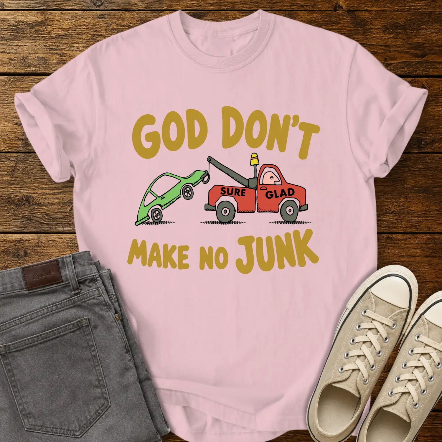 blossom pink T-shirt featuring a humorous cartoon of a red tow truck labeled “SURE GLAD” towing a broken green car beneath the phrase “GOD DON’T MAKE NO JUNK.” The shirt is displayed on a rustic wood plank background with folded gray jeans, cream canvas sneakers, and a casual flat-lay presentation.