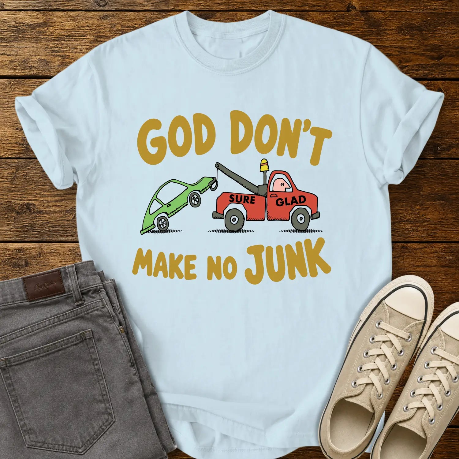 chambray blue T-shirt featuring a humorous cartoon of a red tow truck labeled “SURE GLAD” towing a broken green car beneath the phrase “GOD DON’T MAKE NO JUNK.” The shirt is displayed on a rustic wood plank background with folded gray jeans, cream canvas sneakers, and a casual flat-lay presentation.
