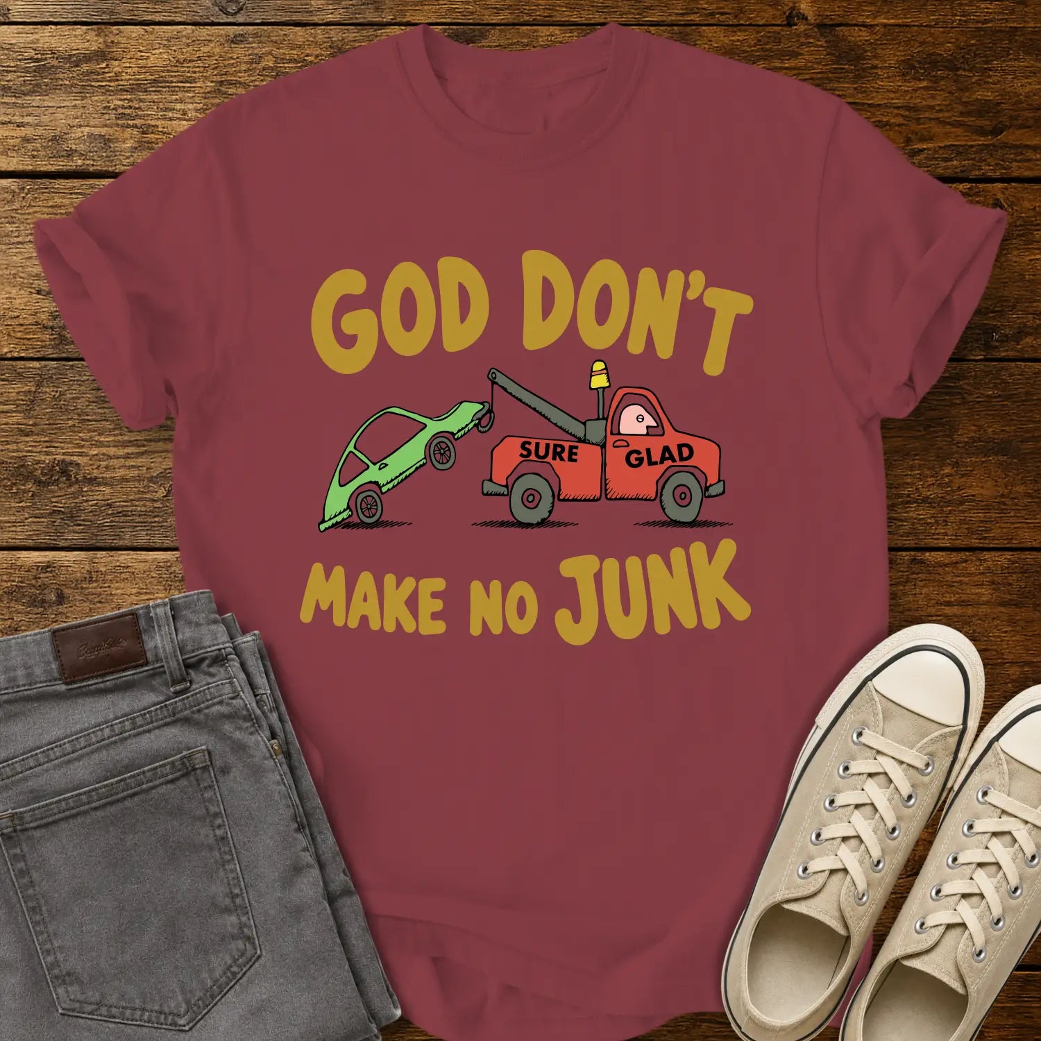 chili red T-shirt featuring a humorous cartoon of a red tow truck labeled “SURE GLAD” towing a broken green car beneath the phrase “GOD DON’T MAKE NO JUNK.” The shirt is displayed on a rustic wood plank background with folded gray jeans, cream canvas sneakers, and a casual flat-lay presentation.