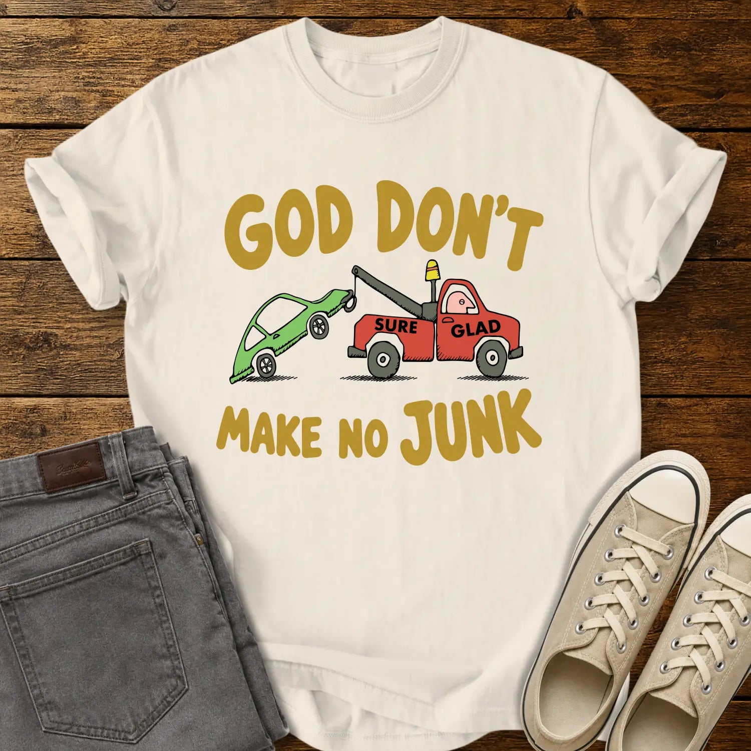 ivory T-shirt featuring a humorous cartoon of a red tow truck labeled “SURE GLAD” towing a broken green car beneath the phrase “GOD DON’T MAKE NO JUNK.” The shirt is displayed on a rustic wood plank background with folded gray jeans, cream canvas sneakers, and a casual flat-lay presentation.