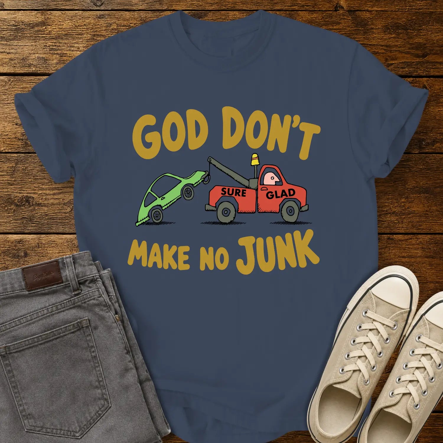 midnight blue T-shirt featuring a humorous cartoon of a red tow truck labeled “SURE GLAD” towing a broken green car beneath the phrase “GOD DON’T MAKE NO JUNK.” The shirt is displayed on a rustic wood plank background with folded gray jeans, cream canvas sneakers, and a casual flat-lay presentation.