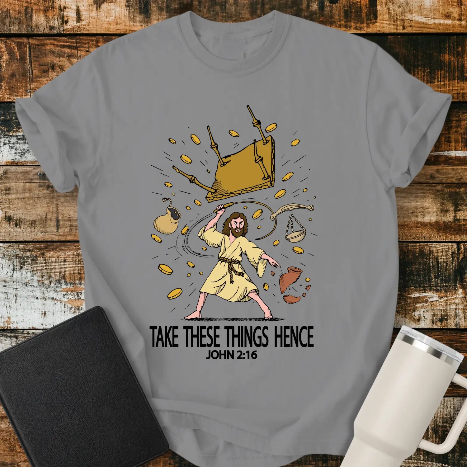 grey graphic T-shirt mockup featuring an illustration of Jesus driving out the money changers, with overturned tables, scattered coins, and the phrase “Take These Things Hence” above a John 2:16 reference, styled with a black Bible and white tumbler on a rustic wood background.