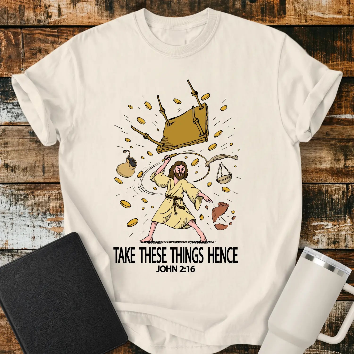 ivory graphic T-shirt mockup featuring an illustration of Jesus driving out the money changers, with overturned tables, scattered coins, and the phrase “Take These Things Hence” above a John 2:16 reference, styled with a black Bible and white tumbler on a rustic wood background.