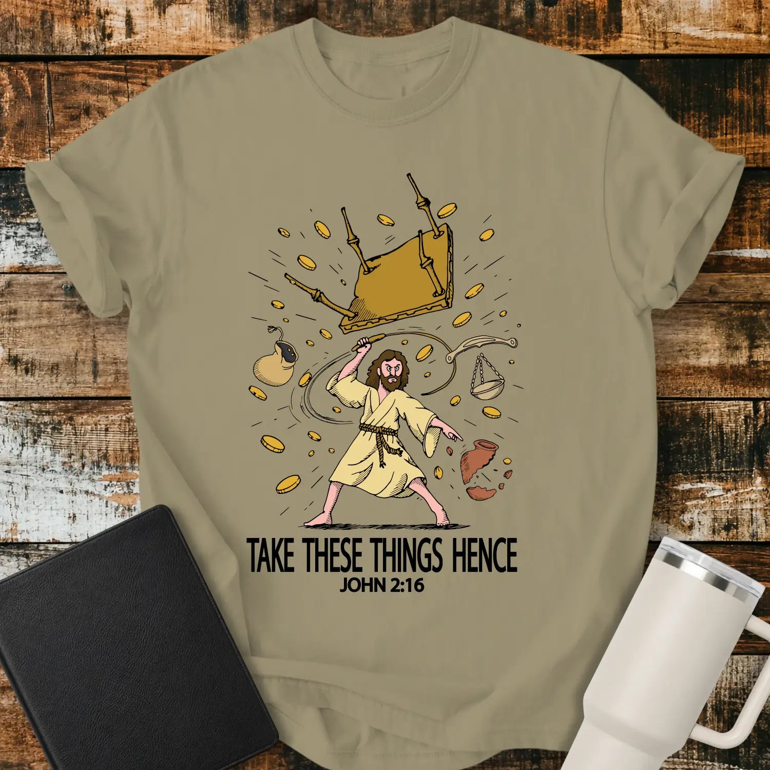 khaki graphic T-shirt mockup featuring an illustration of Jesus driving out the money changers, with overturned tables, scattered coins, and the phrase “Take These Things Hence” above a John 2:16 reference, styled with a black Bible and white tumbler on a rustic wood background.