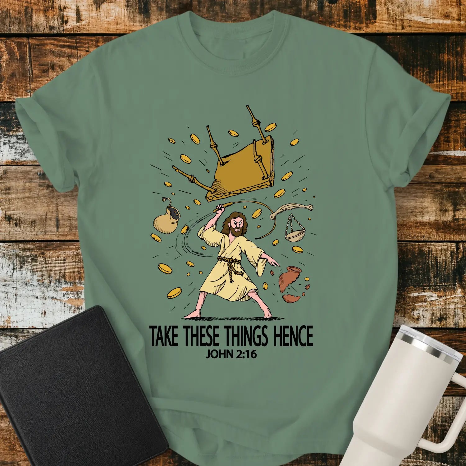light green graphic T-shirt mockup featuring an illustration of Jesus driving out the money changers, with overturned tables, scattered coins, and the phrase “Take These Things Hence” above a John 2:16 reference, styled with a black Bible and white tumbler on a rustic wood background.