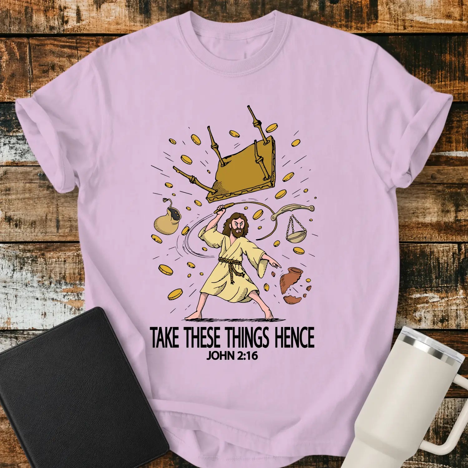 orchid pink graphic T-shirt mockup featuring an illustration of Jesus driving out the money changers, with overturned tables, scattered coins, and the phrase “Take These Things Hence” above a John 2:16 reference, styled with a black Bible and white tumbler on a rustic wood background.