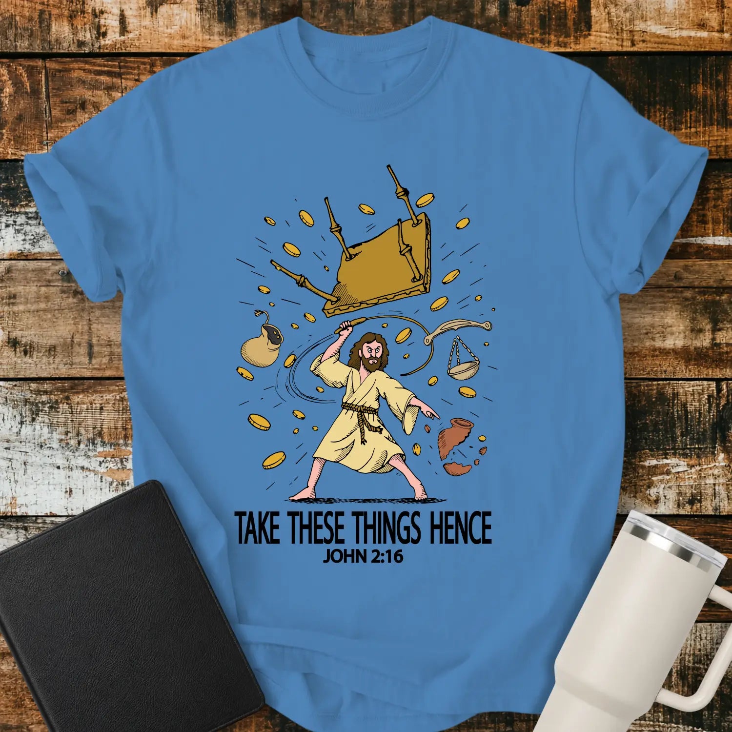 Royal Caribbean blue graphic T-shirt mockup featuring an illustration of Jesus driving out the money changers, with overturned tables, scattered coins, and the phrase “Take These Things Hence” above a John 2:16 reference, styled with a black Bible and white tumbler on a rustic wood background.