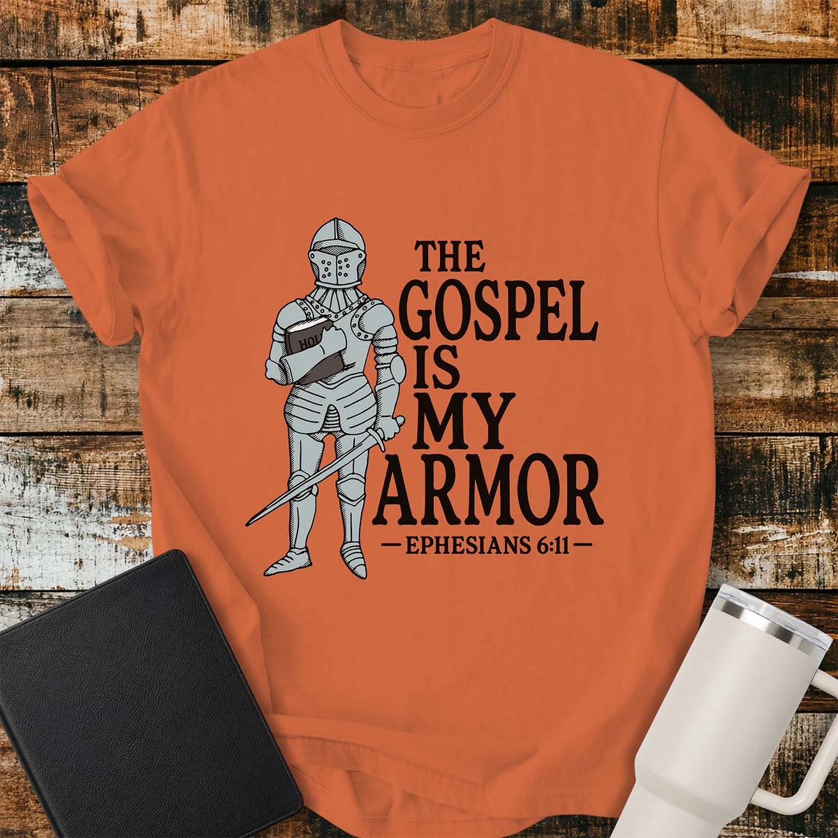 burnt orange flat-lay t-shirt mockup featuring a vintage-style illustration of a medieval knight in full armor holding a Bible and a sword. Bold black lettering reads, "THE GOSPEL IS MY ARMOR," with the Bible reference "EPHESIANS 6:11" below. The shirt is displayed on a rustic wood plank background alongside a black leather Bible and a white insulated travel mug.