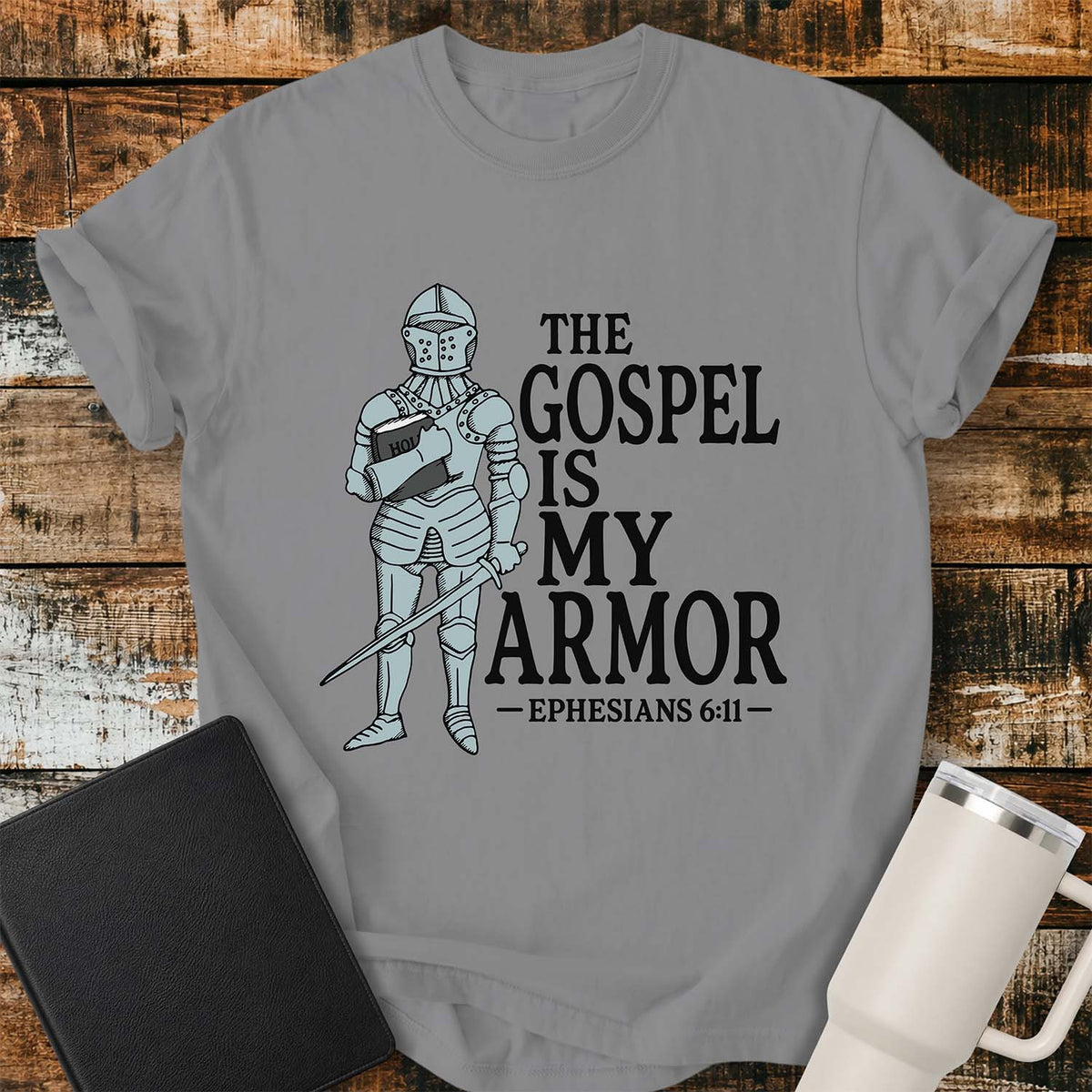 grey flat-lay t-shirt mockup featuring a vintage-style illustration of a medieval knight in full armor holding a Bible and a sword. Bold black lettering reads, "THE GOSPEL IS MY ARMOR," with the Bible reference "EPHESIANS 6:11" below. The shirt is displayed on a rustic wood plank background alongside a black leather Bible and a white insulated travel mug.