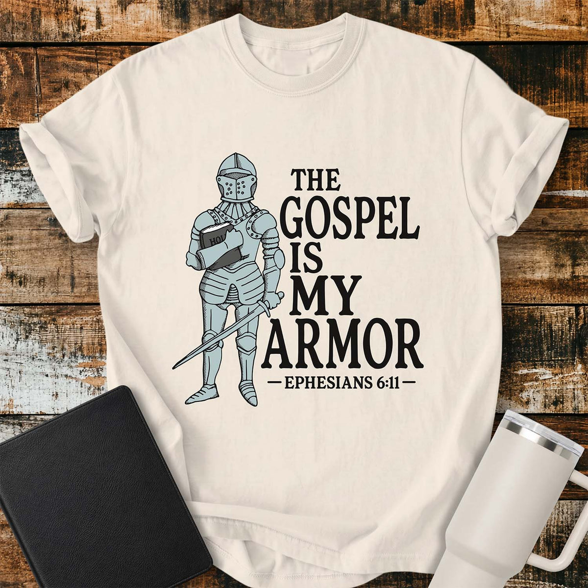ivory flat-lay t-shirt mockup featuring a vintage-style illustration of a medieval knight in full armor holding a Bible and a sword. Bold black lettering reads, "THE GOSPEL IS MY ARMOR," with the Bible reference "EPHESIANS 6:11" below. The shirt is displayed on a rustic wood plank background alongside a black leather Bible and a white insulated travel mug.
