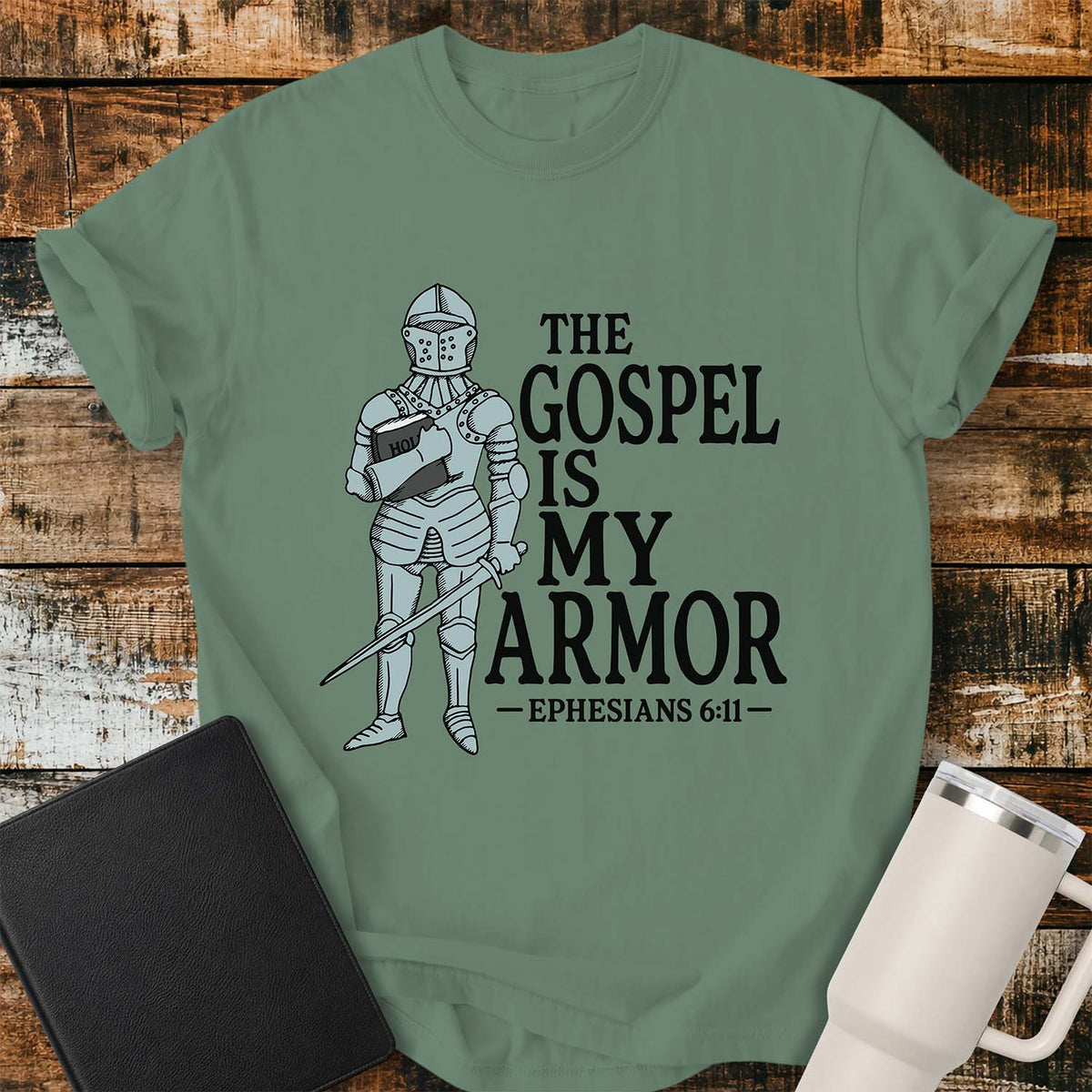 light green flat-lay t-shirt mockup featuring a vintage-style illustration of a medieval knight in full armor holding a Bible and a sword. Bold black lettering reads, "THE GOSPEL IS MY ARMOR," with the Bible reference "EPHESIANS 6:11" below. The shirt is displayed on a rustic wood plank background alongside a black leather Bible and a white insulated travel mug.