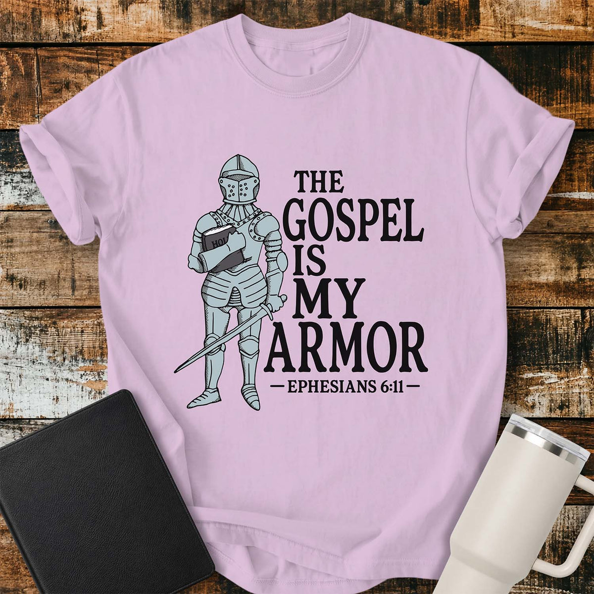orchid pink flat-lay t-shirt mockup featuring a vintage-style illustration of a medieval knight in full armor holding a Bible and a sword. Bold black lettering reads, "THE GOSPEL IS MY ARMOR," with the Bible reference "EPHESIANS 6:11" below. The shirt is displayed on a rustic wood plank background alongside a black leather Bible and a white insulated travel mug.