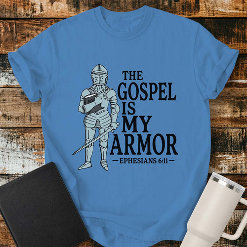 Royal caribe blue flat-lay t-shirt mockup featuring a vintage-style illustration of a medieval knight in full armor holding a Bible and a sword. Bold black lettering reads, "THE GOSPEL IS MY ARMOR," with the Bible reference "EPHESIANS 6:11" below. The shirt is displayed on a rustic wood plank background alongside a black leather Bible and a white insulated travel mug.